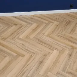 Ułożona pod kątem jasna podłoga z paneli imitujących drewno, widoczne białe listwy przypodłogowe, ściana w kolorze granatowym i szarym, gniazdka elektryczne oraz przewody przy ścianie po lewej...