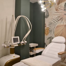 Book Your Beauty SC - Jasny gabinet z białym fotelem zabiegowym przykrytym pledem, nowoczesnym urządzeniem kosmetycznym i złotym żyrandolem na tle ściany z motywem roślinnym.