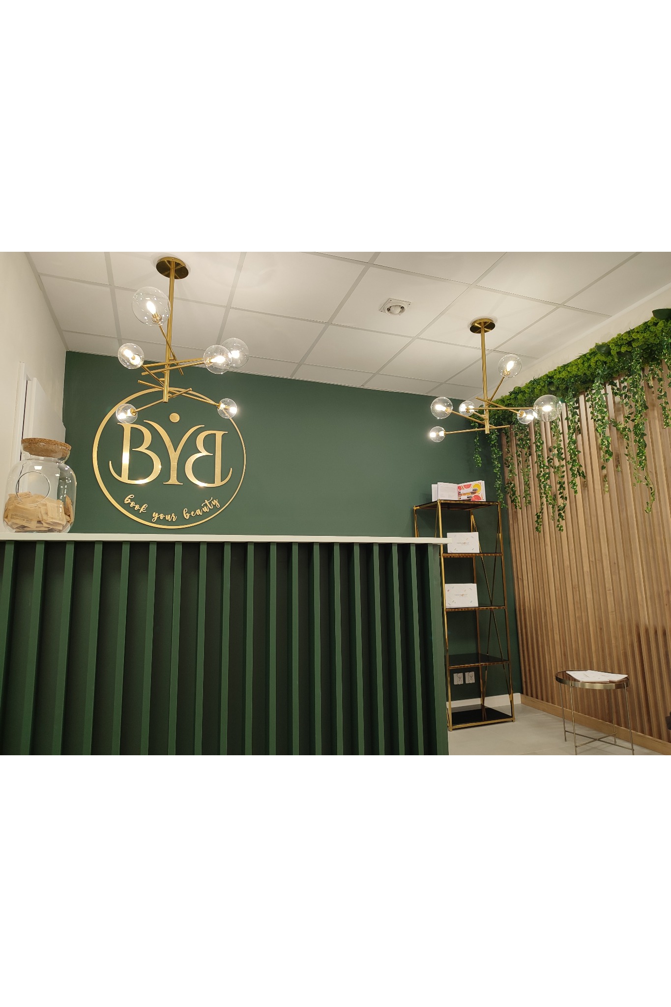 Recepcja w salonie kosmetycznym z zielonymi ścianami, złotym logo 'BYB Book your beauty' i nowoczesnym oświetleniem.