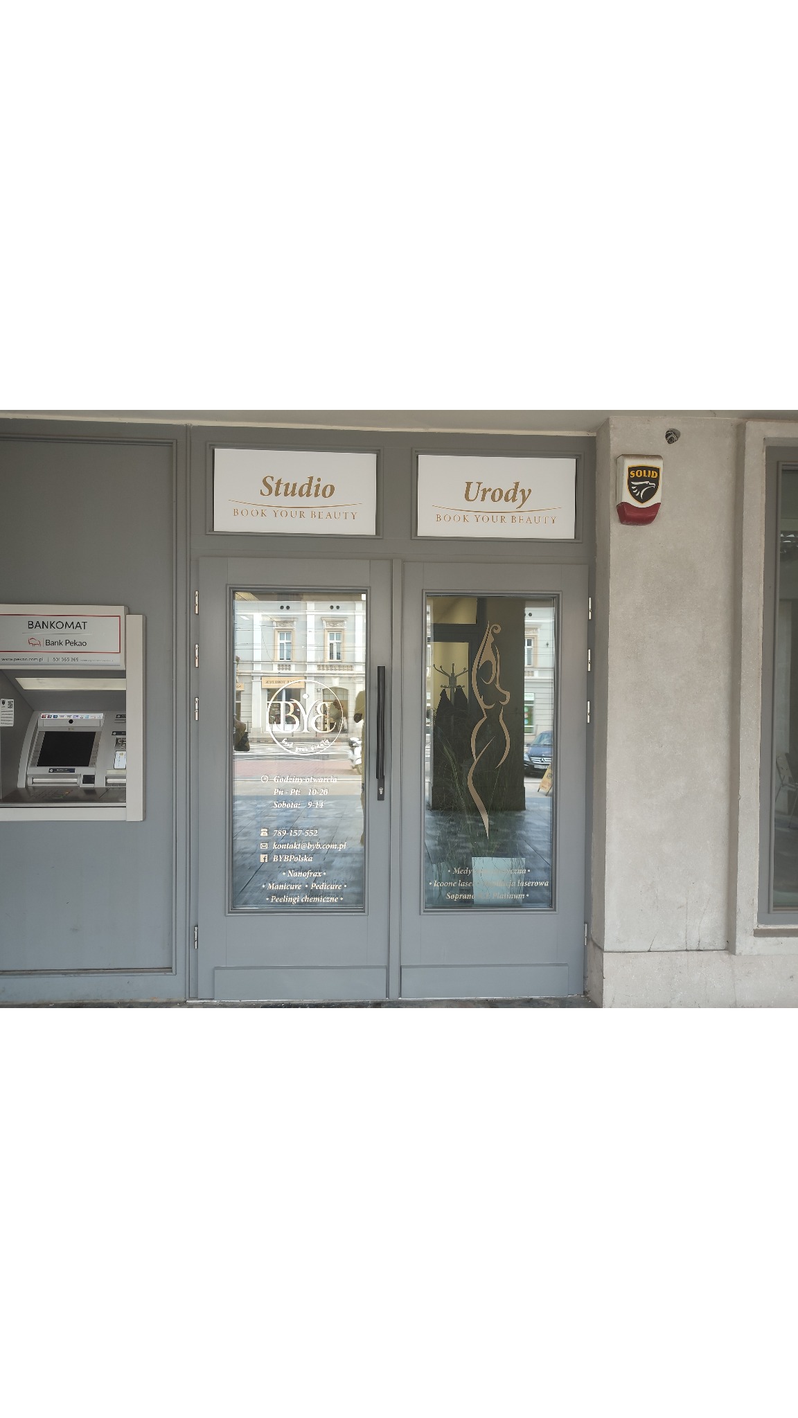Wejście do Studio Urody 'Book Your Beauty' z szarymi drzwiami i witrynami, widoczne godziny otwarcia, logo firmy na szybie i lista oferowanych usług, w tle ulica miejska.
