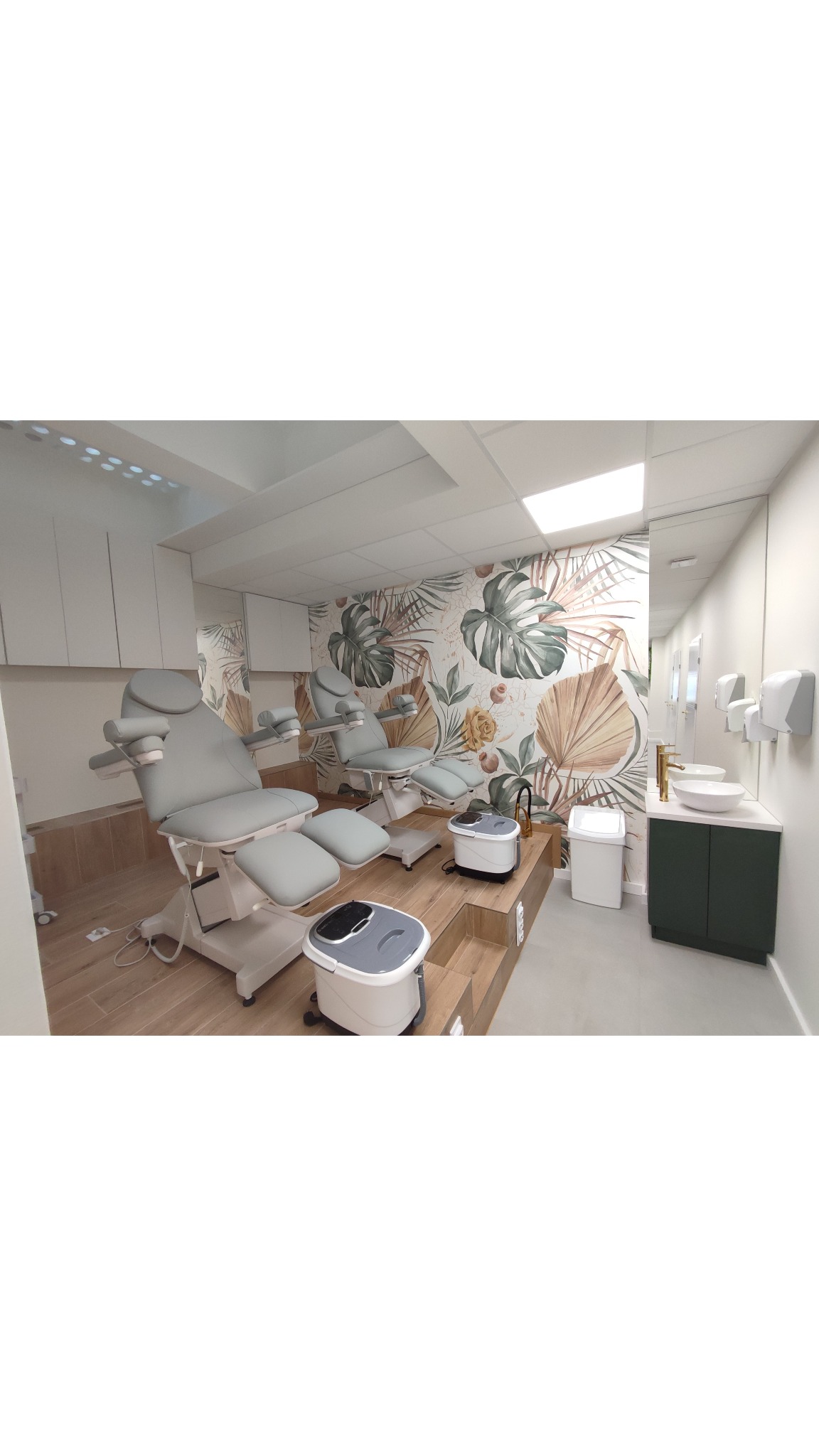 Jasne wnętrze gabinetu z dwoma fotelami do pedicure, urządzeniami do hydromasażu stóp, umywalką z zieloną szafką i tapetą w liście.