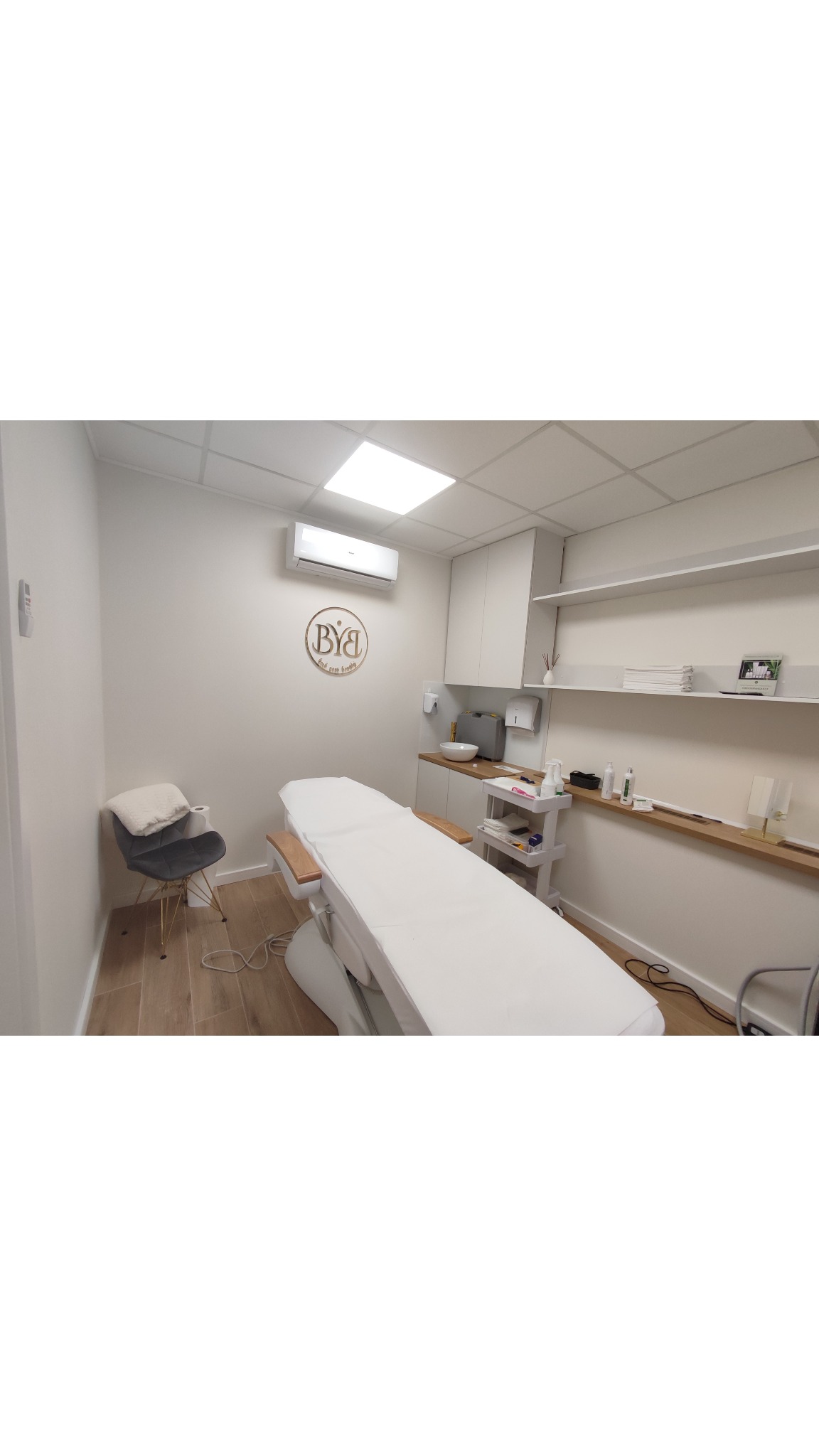 Jasne, minimalistyczne wnętrze gabinetu z regulowanym łóżkiem kosmetycznym, krzesłem dla klienta i umywalką z akcesoriami na półkach.