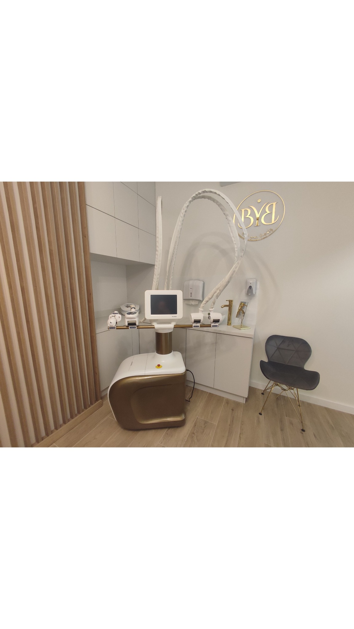 Nowoczesny gabinet z urządzeniem do modelowania sylwetki w kolorze białym i złotym, obok szara krzesło na złotych nóżkach, w tle logo firmy na ścianie.