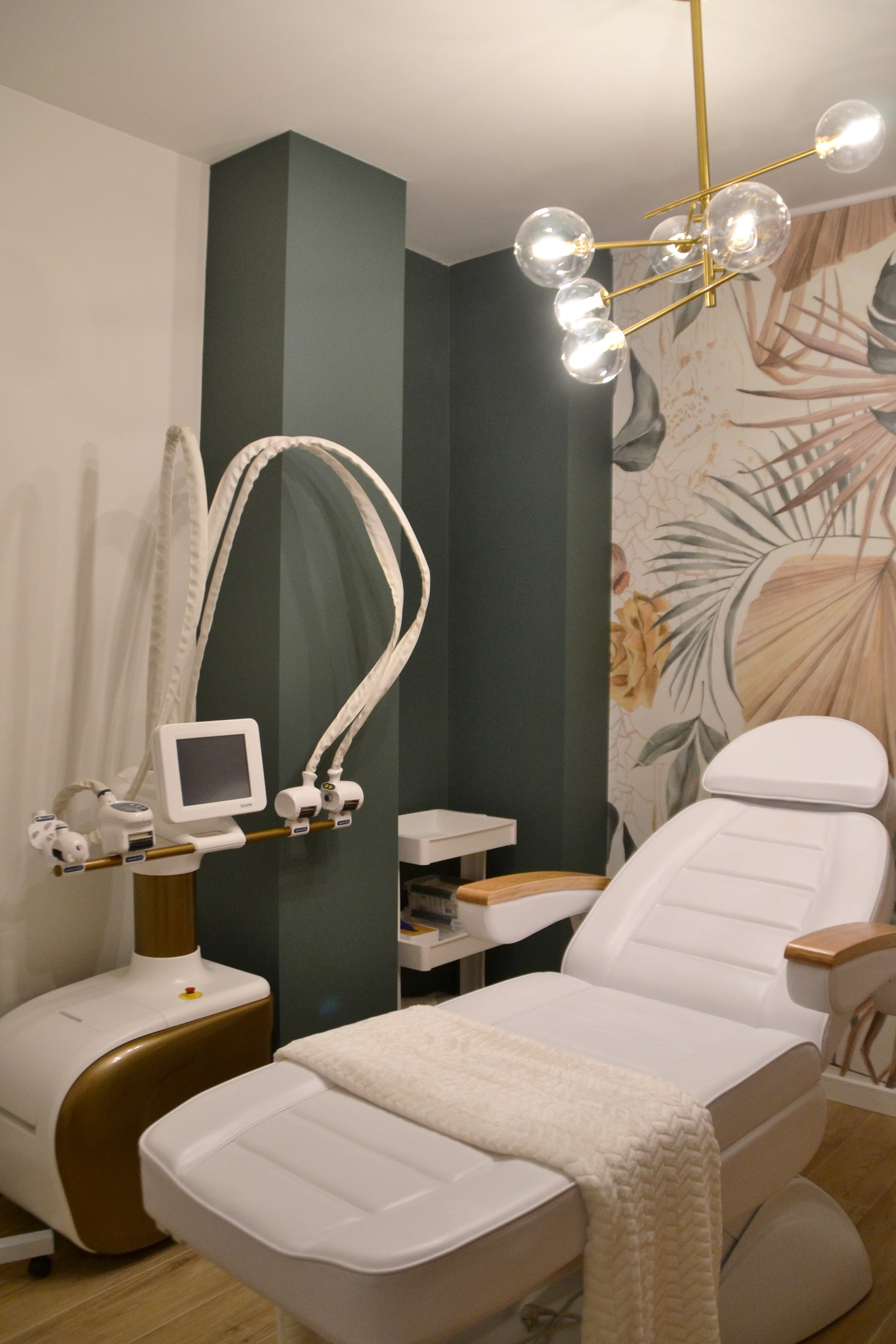 Jasny gabinet z białym fotelem zabiegowym przykrytym pledem, nowoczesnym urządzeniem kosmetycznym i złotym żyrandolem na tle ściany z motywem roślinnym.