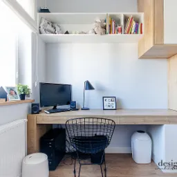 Nowoczesne biurko z drewnianym blatem i białymi szafkami, czarne krzesło druciane, komputer i lampka na biurku, kalendarz i ramka ze zdjęciem na parapecie okna, półka z książkami i dekoracjami...