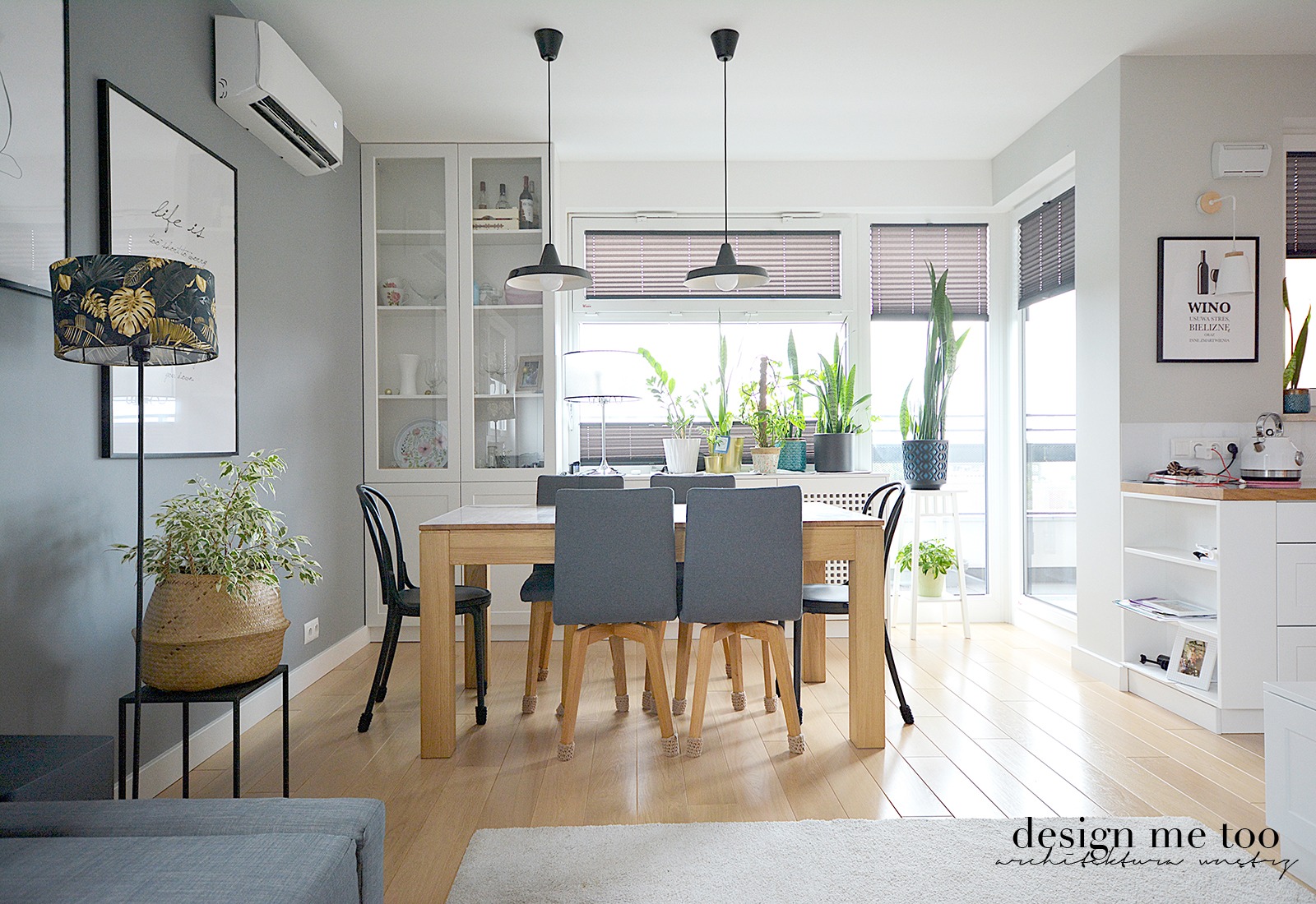 Jasne, nowoczesne wnętrze jadalni z drewnianym stołem otoczonym szarymi krzesłami, oświetlone dwoma czarnymi lampami wiszącymi. W tle okno z żaluzjami, liczne rośliny doniczkowe i biała zabudowa...