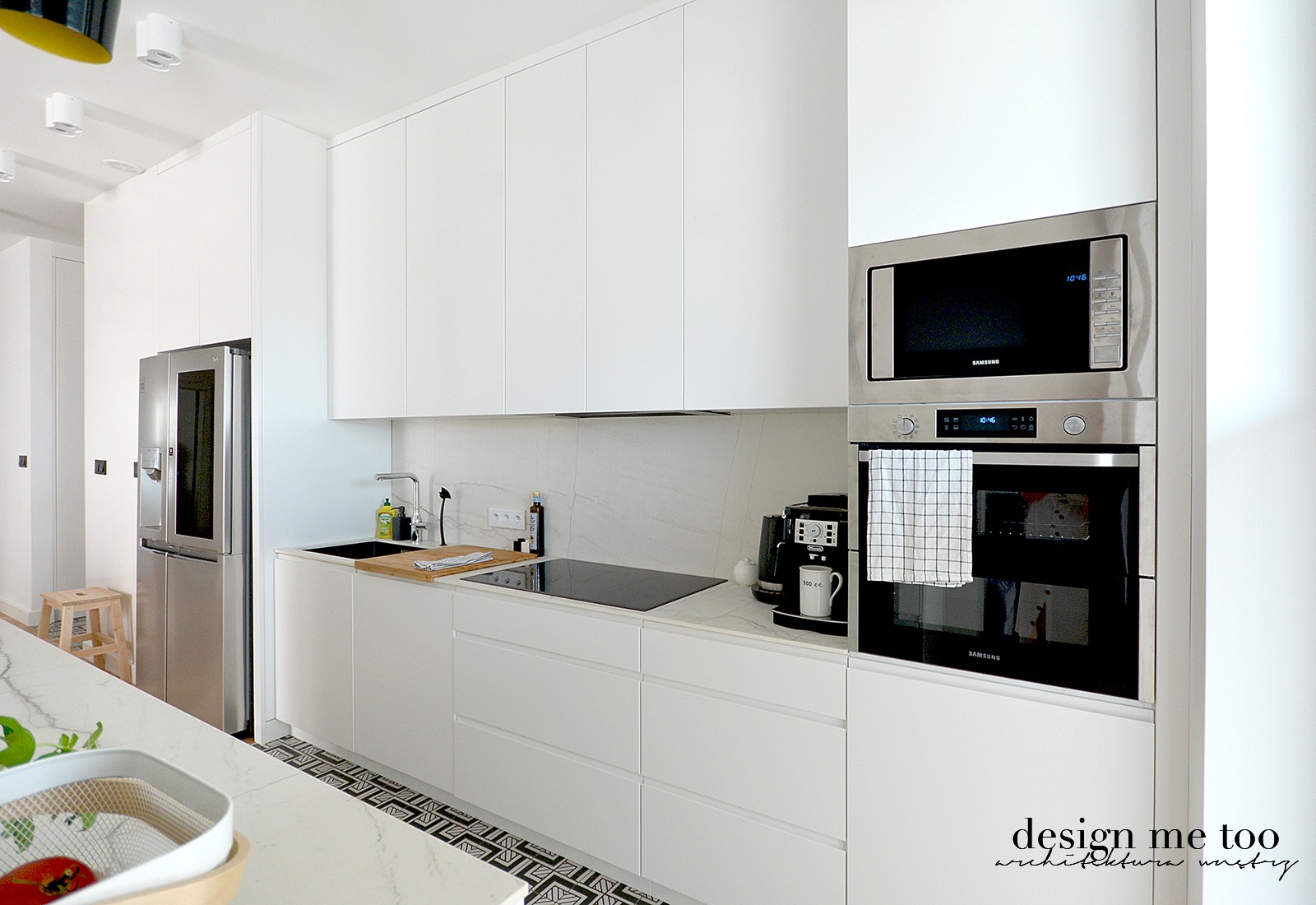 Minimalistyczna kuchnia w bieli: zabudowa kuchenna z piekarnikiem i mikrofalówką Samsung, ekspres do kawy Delonghi, czarny zlew, marmurowy blat i wzorzysta podłoga.