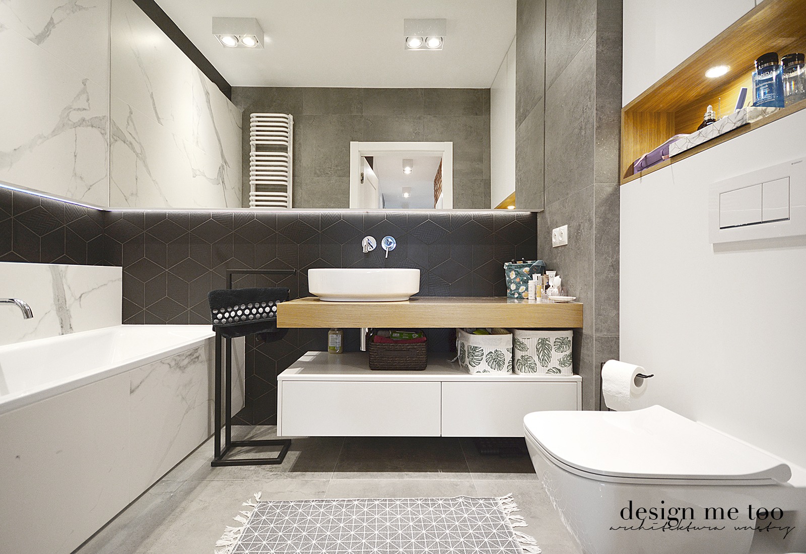 Nowoczesna łazienka z marmurową wanną, umywalką nablatową na drewnianym blacie, szarą mozaiką heksagonalną i białymi szafkami pod umywalką, uzupełniona o geometryczny dywanik i dekoracyjne kosze...