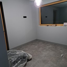 Usługi Wykończeniowe Tomasz Krawczyński - Pokój po remoncie: szare ściany, szara podłoga z paneli, okno z drewnianą ramą, żarówka na kablu, zakryte folią przedmioty w rogu.