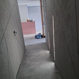 Usługi Wykończeniowe Tomasz Krawczyński - Korytarz w trakcie wykańczania z szarymi panelami ściennymi o geometrycznym wzorze i wbudowanym oświetleniem LED, widoczne materiały budowlane i narzędzia w tle.