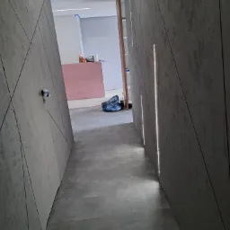 Korytarz w trakcie wykańczania z szarymi panelami ściennymi o geometrycznym wzorze i wbudowanym oświetleniem LED, widoczne materiały budowlane i narzędzia w tle.