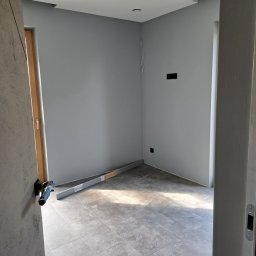 Usługi Wykończeniowe Tomasz Krawczyński - Szare płytki podłogowe imitujące kamień w nowym pomieszczeniu, z poziomnicą przy ścianie i widocznymi puszkami elektrycznymi, ściany pomalowane na szaro, widok zza futryny.