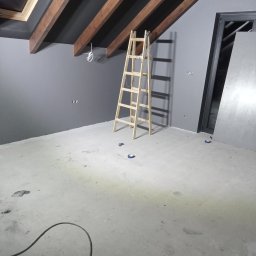 Usługi Wykończeniowe Tomasz Krawczyński - Pokój w Nowym Mieście nad Wartą w trakcie prac wykończeniowych, widoczne szare ściany, drewniane belki stropowe, surowe podłoże, drabina i zabezpieczone gniazdka elektryczne.