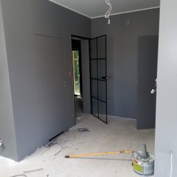 Usługi Wykończeniowe Tomasz Krawczyński - Wnętrze pomalowane na szaro z czarnymi drzwiami w stylu industrialnym, widoczny wałek do malowania i puszka farby, surowy beton na podłodze, przygotowanie do dalszych prac wykończeniowych.