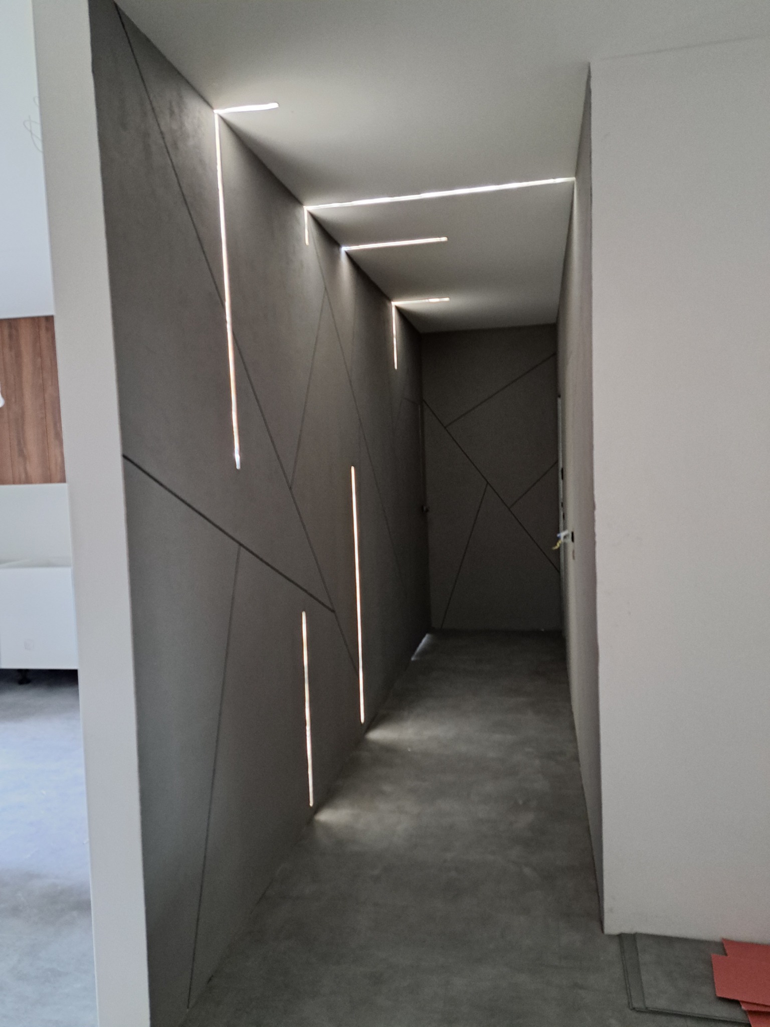 Nowoczesny korytarz z szarą ścianą o geometrycznym wzorze i wbudowanym oświetleniem LED, surowa betonowa podłoga, widok z perspektywy wejścia.
