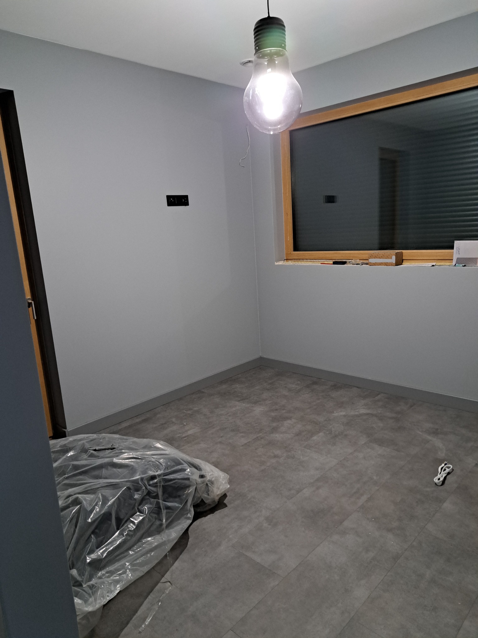 Pokój po remoncie: szare ściany, szara podłoga z paneli, okno z drewnianą ramą, żarówka na kablu, zakryte folią przedmioty w rogu.