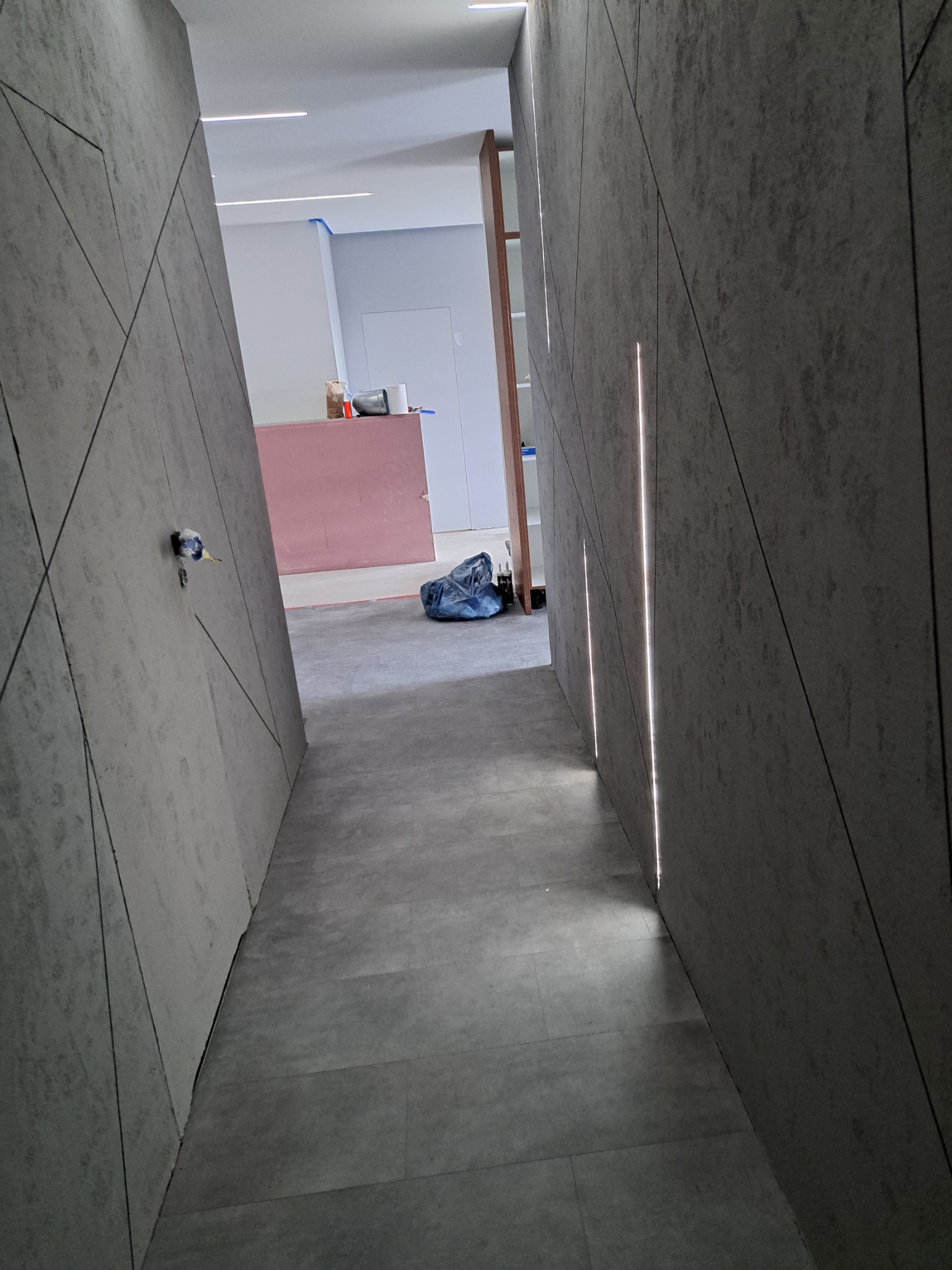 Korytarz w trakcie wykańczania z szarymi panelami ściennymi o geometrycznym wzorze i wbudowanym oświetleniem LED, widoczne materiały budowlane i narzędzia w tle.
