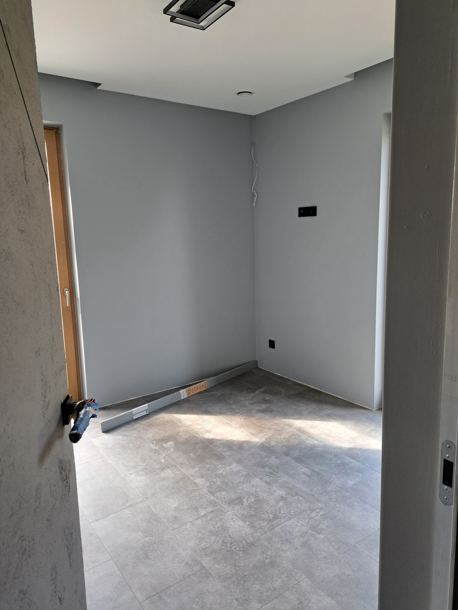 Szare płytki podłogowe imitujące kamień w nowym pomieszczeniu, z poziomnicą przy ścianie i widocznymi puszkami elektrycznymi, ściany pomalowane na szaro, widok zza futryny.