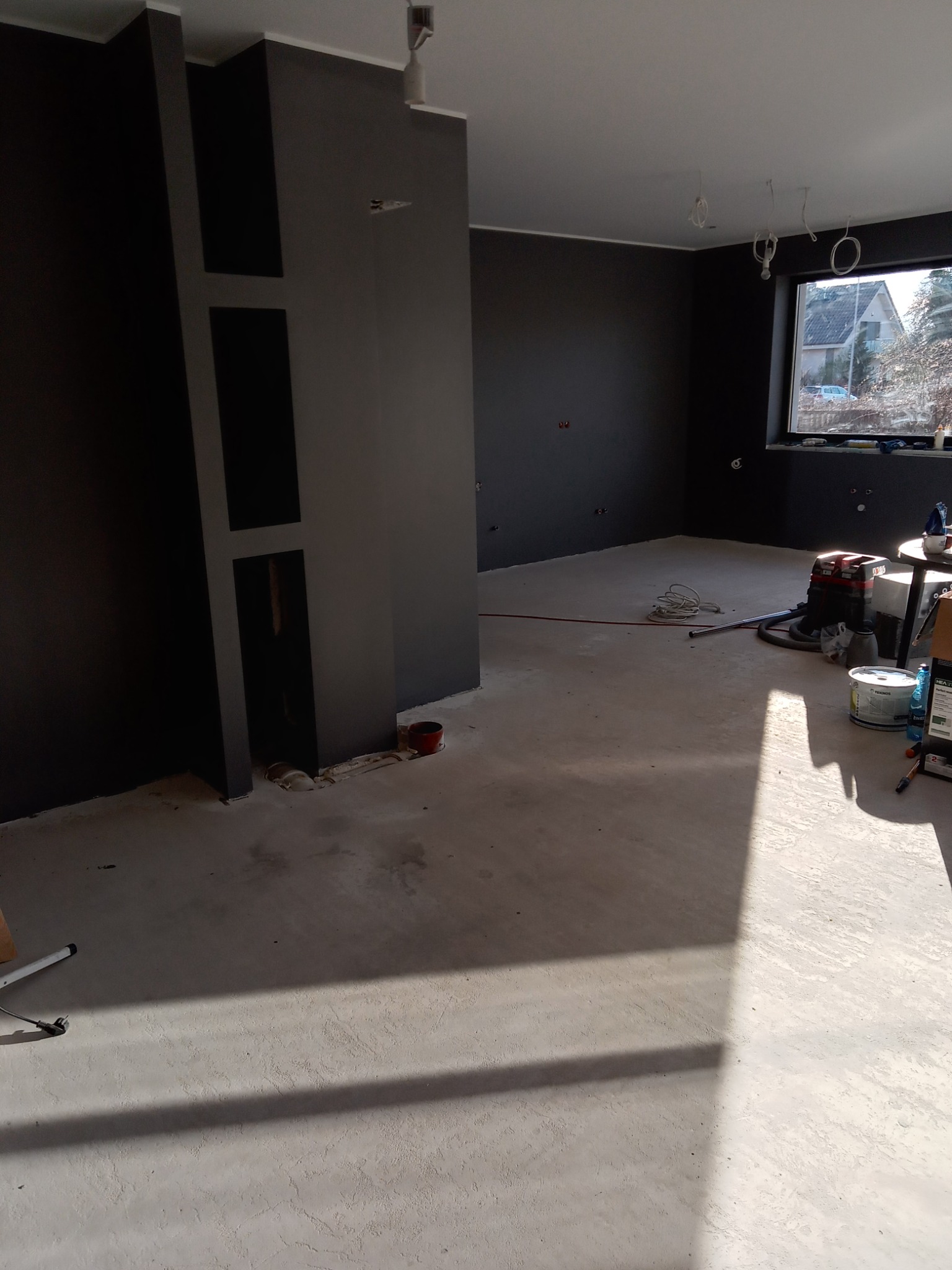 Pokój w trakcie remontu, ściany pomalowane na szaro, widoczne niedoskonałości i przygotowania do dalszych prac wykończeniowych, narzędzia i materiały budowlane na podłodze, wpadające światło...