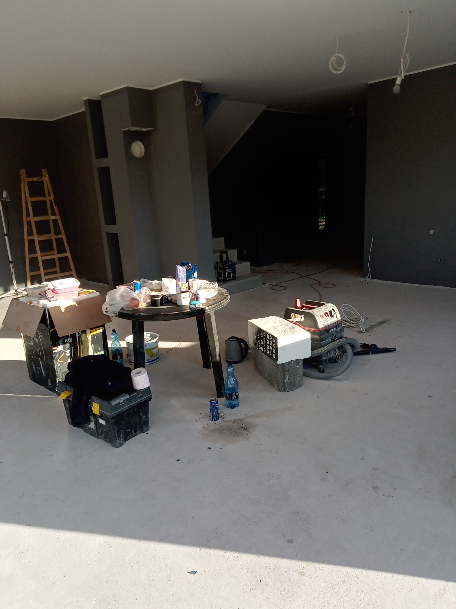 Przygotowania do malowania wnętrza: pomalowane na szaro ściany, drabina oparta o ścianę, stół z przekąskami i napojami, narzędzia i odkurzacz na podłodze, w tle schody.