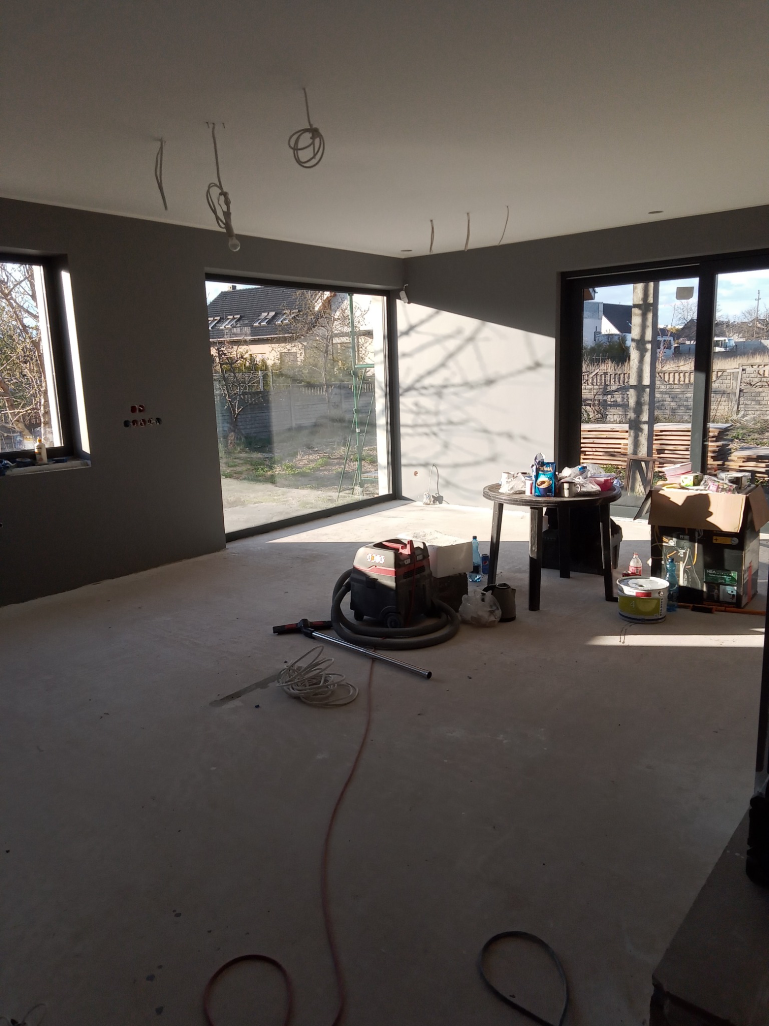 Wnętrze w trakcie przygotowania do prac wykończeniowych: szare ściany, surowy beton na podłodze, widoczne przewody elektryczne, okna z widokiem na ogród, odkurzacz przemysłowy i stół z akcesoriami...