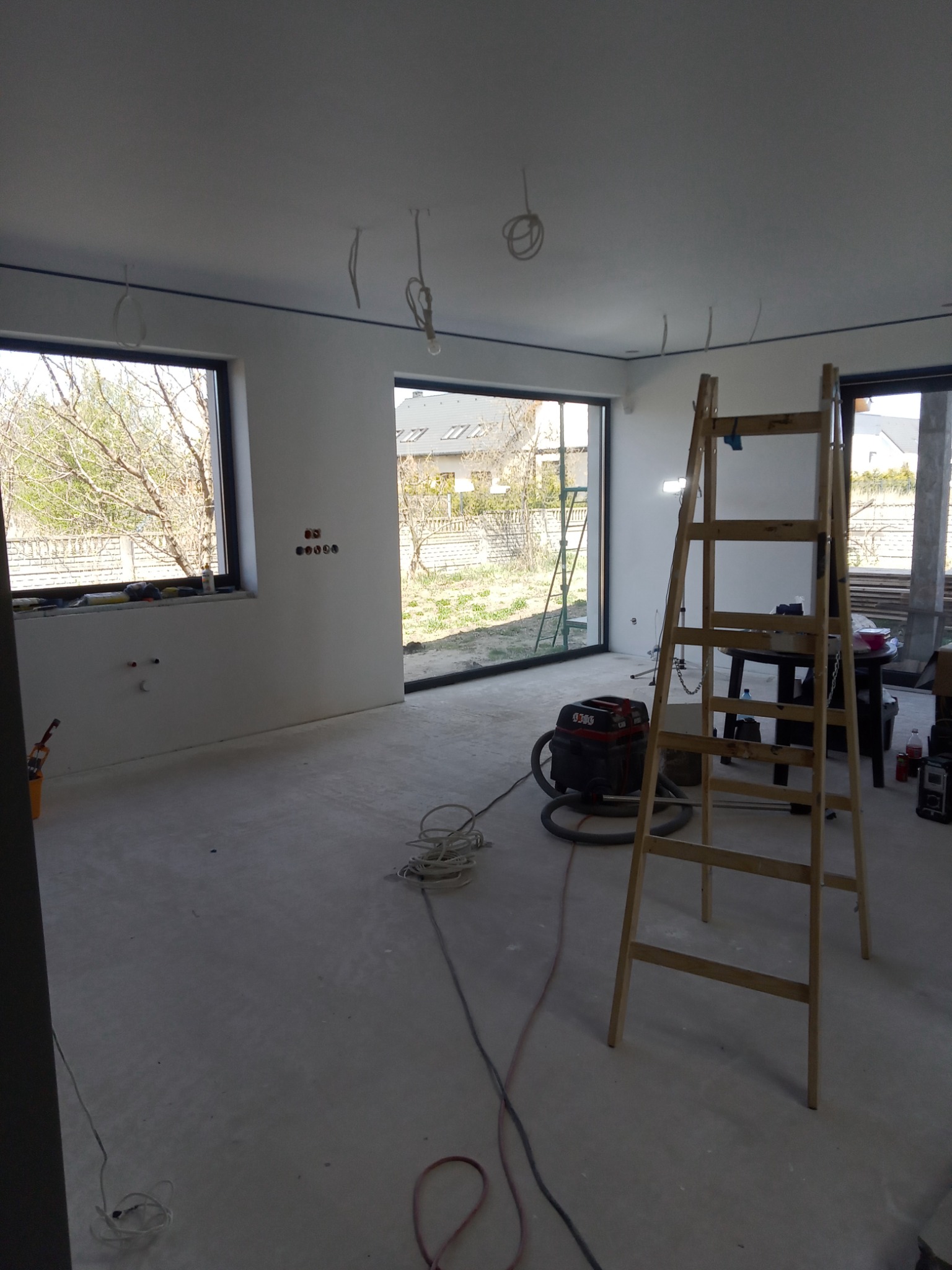 Pokój w trakcie remontu: gładkie, białe ściany, surowa podłoga, drabina, odkurzacz przemysłowy i wystające kable elektryczne.