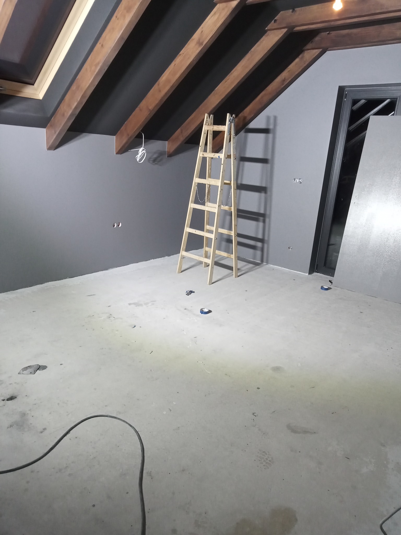 Pokój w Nowym Mieście nad Wartą w trakcie prac wykończeniowych, widoczne szare ściany, drewniane belki stropowe, surowe podłoże, drabina i zabezpieczone gniazdka elektryczne.