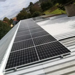 Instalacja paneli słonecznych na dachu z blachy trapezowej; widok z góry na panele w czarnych ramach, mocowane do białej konstrukcji nośnej, z budynkami i drzewami w tle.