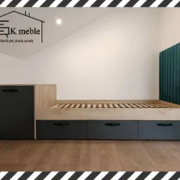 Łóżko z drewnianą ramą i szarymi szufladami, tapicerowane wezgłowie w odcieniach zieleni, widoczne logo firmy meblarskiej w rogu pokoju na poddaszu.