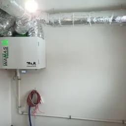 Biała jednostka rekuperacji WANAS 415 HI-TECH z podłączonymi srebrnymi rurami wentylacyjnymi, zamontowana na białej ścianie w trakcie instalacji, widoczne przewody i fragment surowej ściany z cegły.