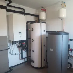 Instalacja pompy ciepła Panasonic Aquarea z dwoma zbiornikami buforowymi w kotłowni w Wieliczce, widoczne czarne rury instalacyjne i białe zbiorniki wyrównawcze.