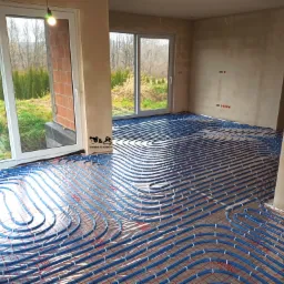 Niebieskie rury ogrzewania podłogowego ułożone na folii z czerwoną kratką w surowym wnętrzu z widokiem na zielony ogród przez duże okna.