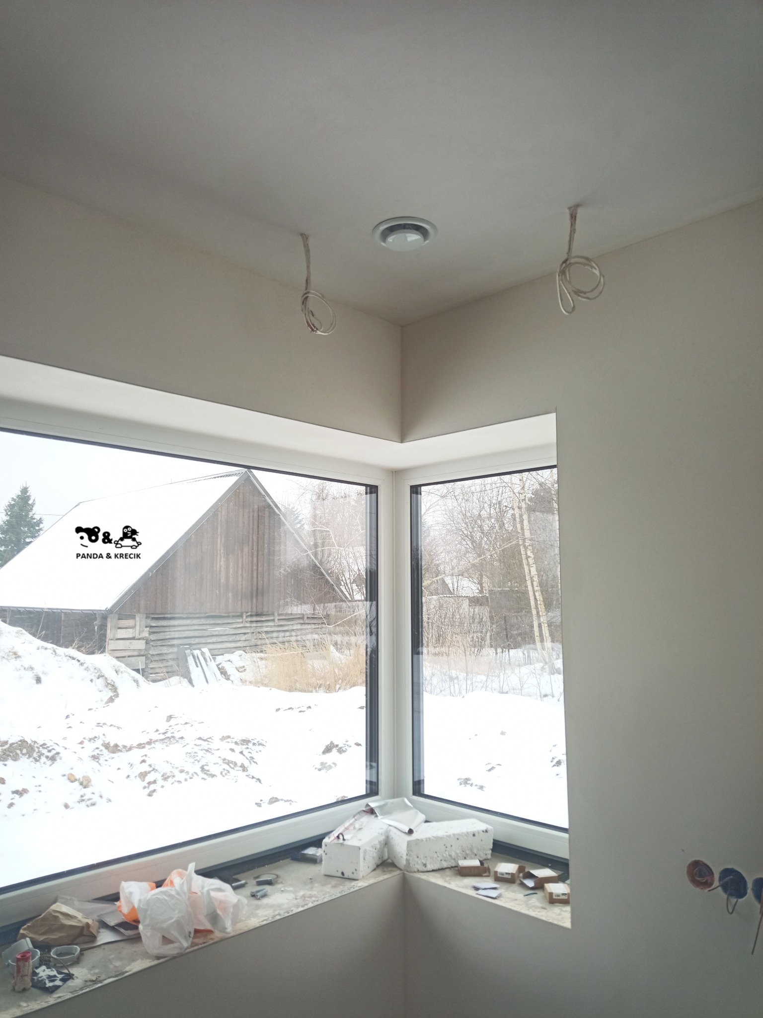 Wnętrze w trakcie wykończenia z dużym narożnym oknem z widokiem na zaśnieżony krajobraz i drewniany budynek gospodarczy, widoczne wystające przewody z sufitu i puszki elektryczne w ścianie,...
