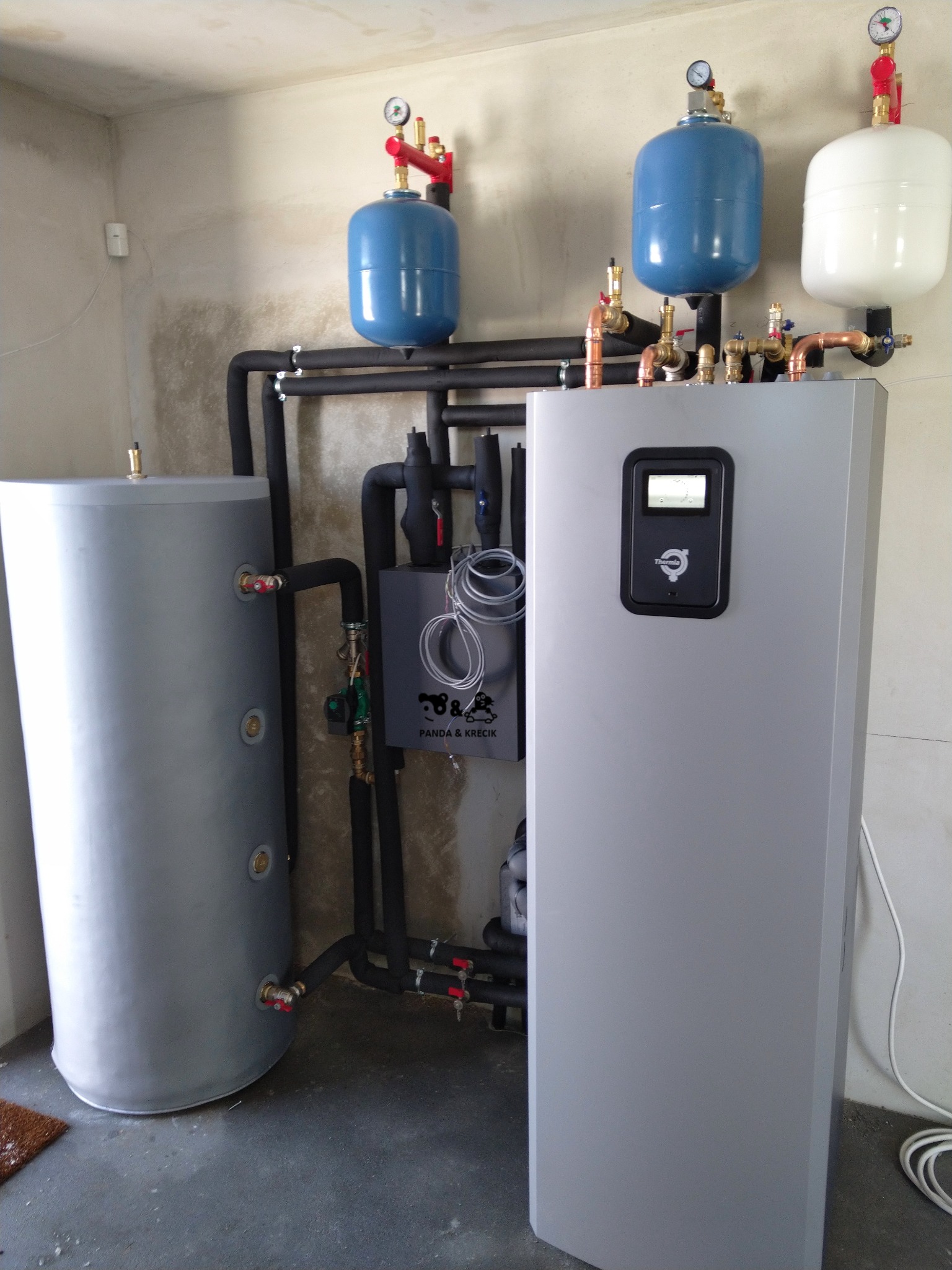Szara instalacja grzewcza z zbiornikiem buforowym i osprzętem, widoczne rury, manometry i logo producenta na urządzeniu.