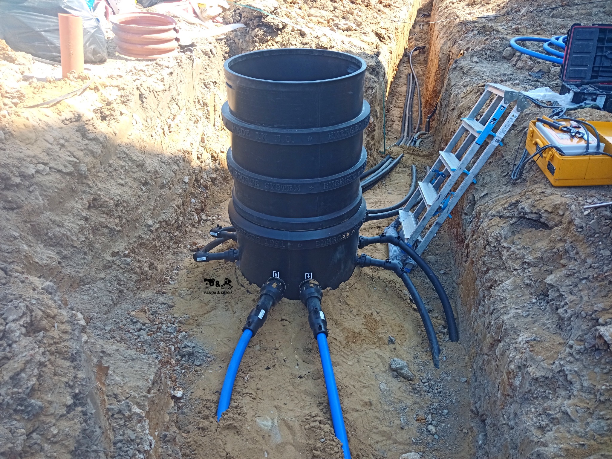 Czarna studzienka rozsączająca w wykopie ziemnym, z podłączonymi niebieskimi rurami, widoczna drabina i skrzynka narzędziowa w tle.