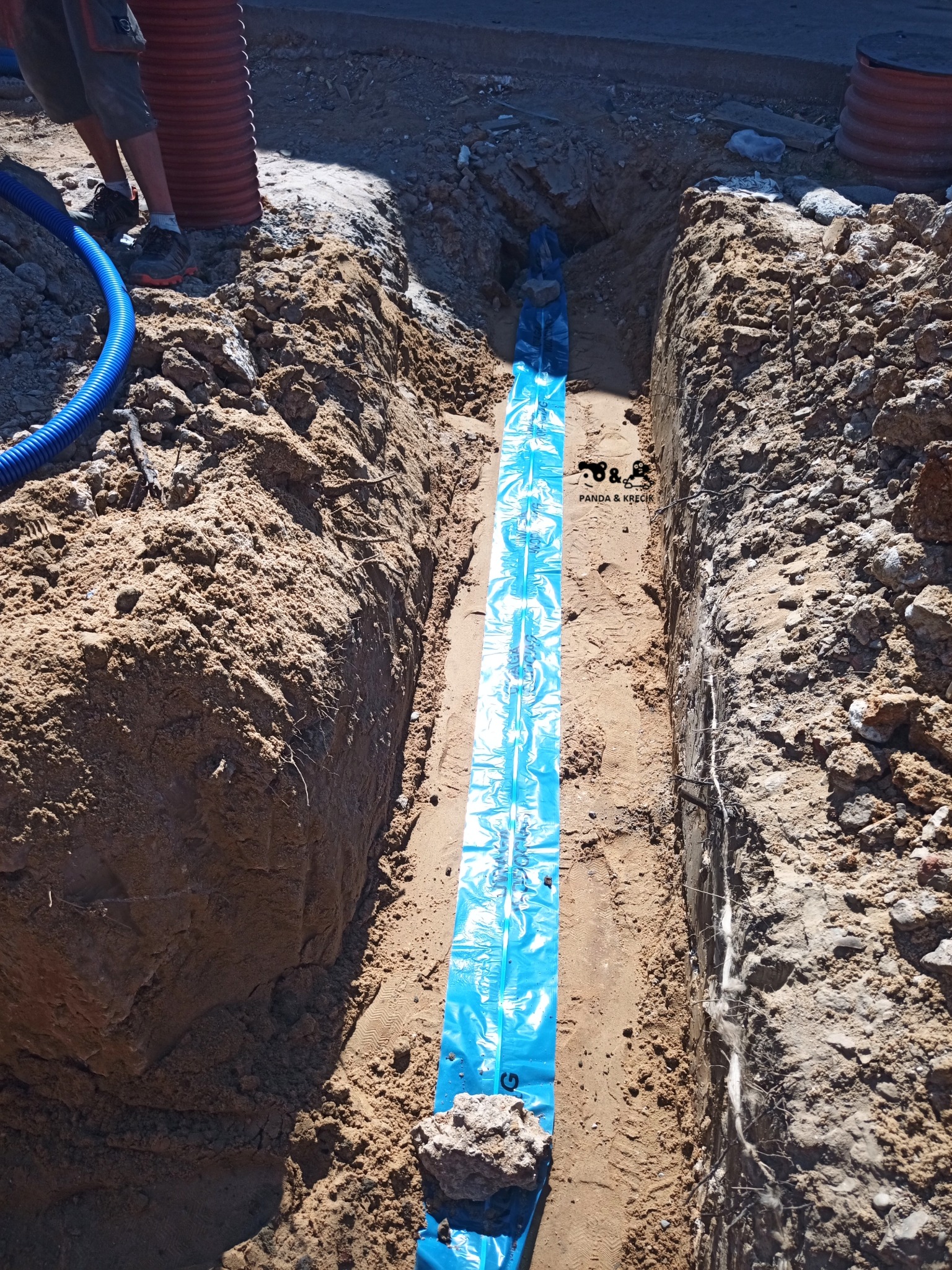 Wykop w ziemi zabezpieczony niebieską folią, prawdopodobnie pod instalację, widoczne rury i fragment osoby w tle.