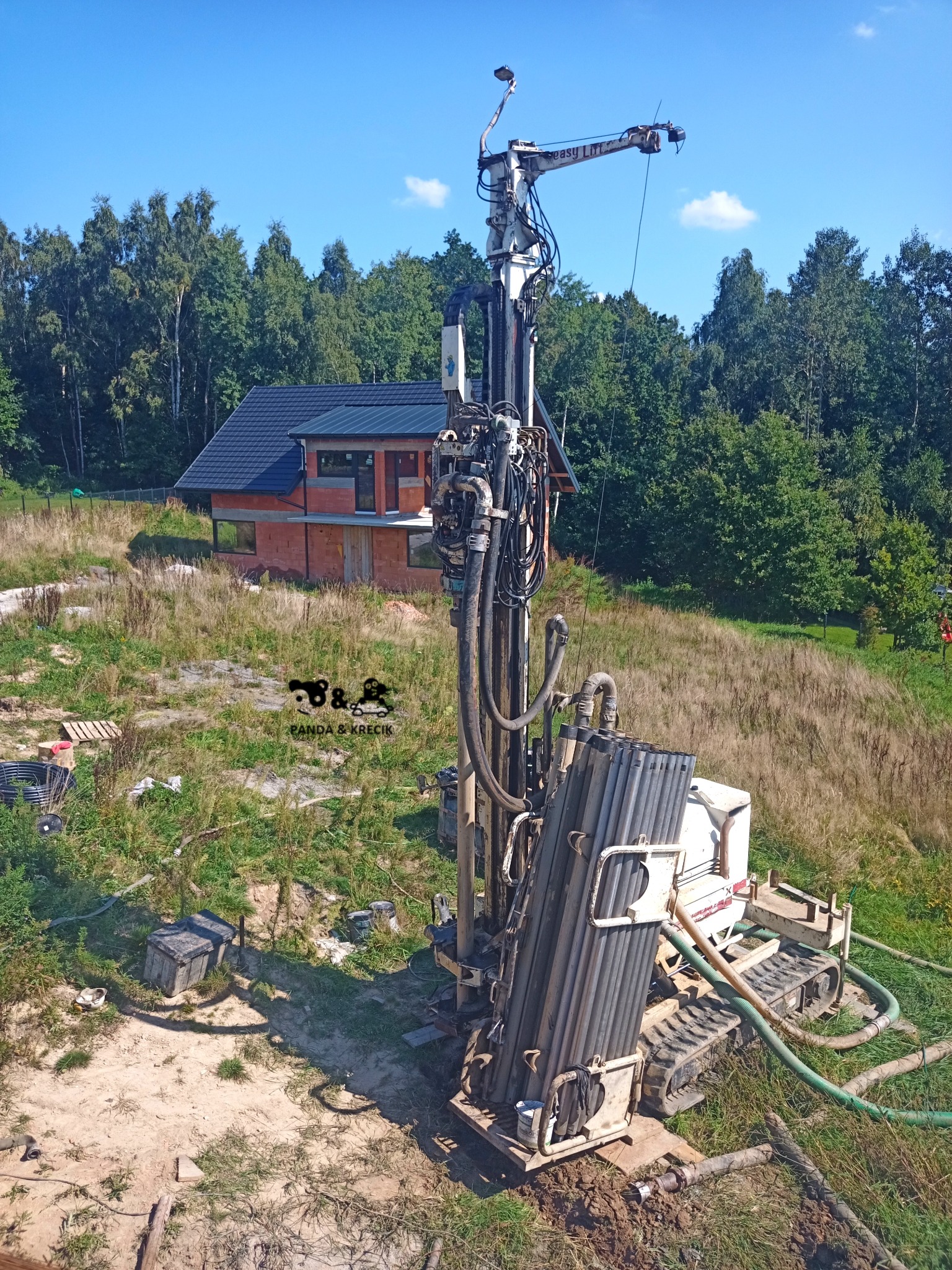 Wiertnica geologiczna, widok z góry, rury wiertnicze, budynek w stanie surowym otwartym w tle, teren trawiasty.
