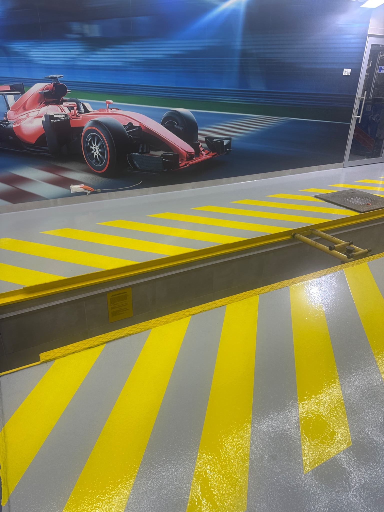 Szaro-żółta podłoga z pasami w garażu w Warszawie, z dekoracyjnym motywem bolidu Formuły 1 na ścianie w tle.