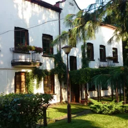 Biały budynek z ciemnymi oknami, porośnięty bluszczem, z małymi balkonami i kwiatami, otoczony zielenią, sfotografowany w słoneczny dzień.