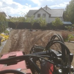 Michał Wawrzyniak - Widok z kabiny traktora na wyrównywany teren działki pod przyszły ogród, z widocznymi śladami opon, domem w tle i przygotowanymi materiałami budowlanymi.