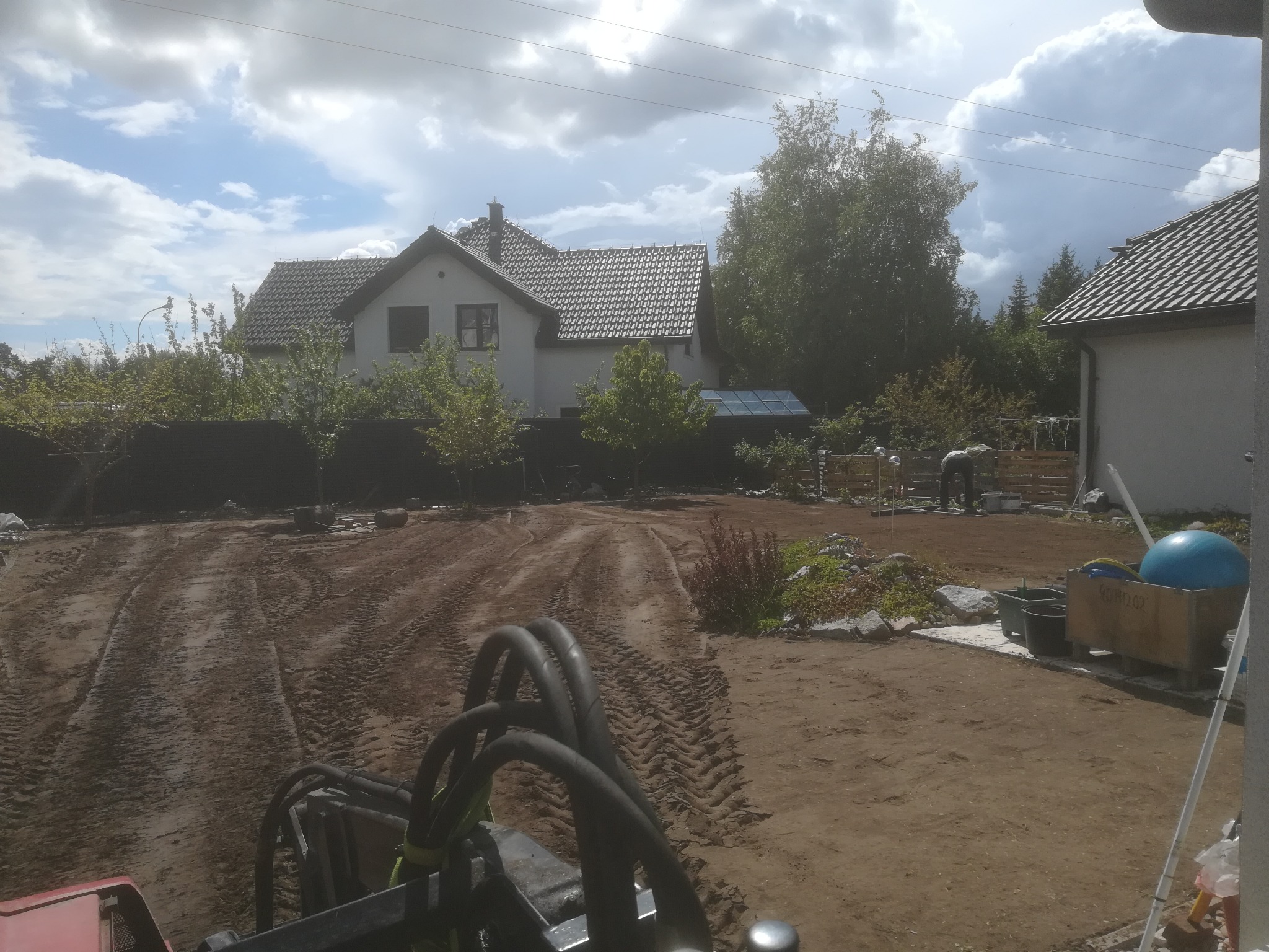 Przygotowany teren pod trawnik z widocznymi śladami opon, częściowo wyrównana ziemia, w tle dom jednorodzinny i pracująca osoba.