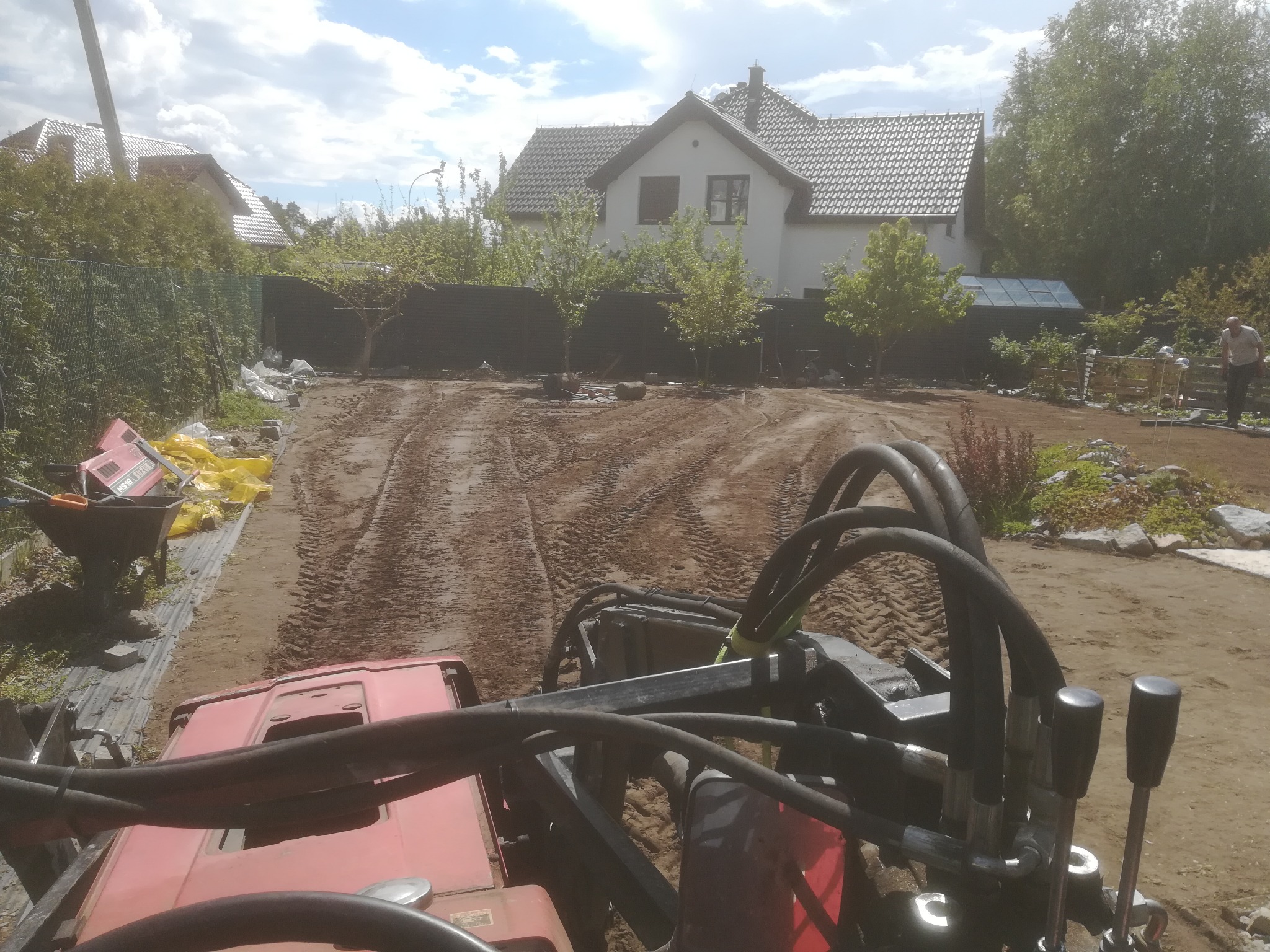 Widok z kabiny traktora na wyrównywany teren działki pod przyszły ogród, z widocznymi śladami opon, domem w tle i przygotowanymi materiałami budowlanymi.