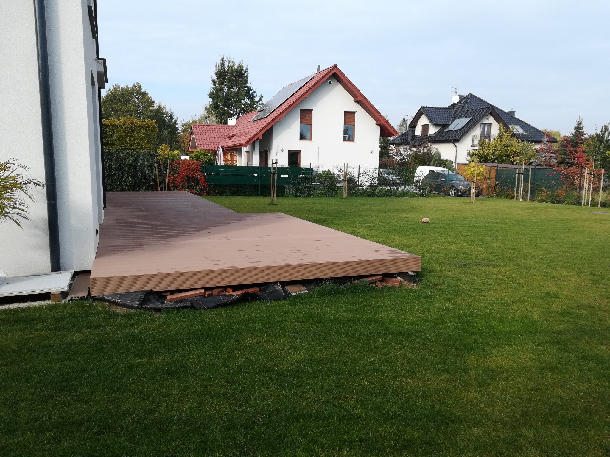 Nowo wybudowany taras kompozytowy przylegający do białego domu, widok na zadbany trawnik i domy w tle.