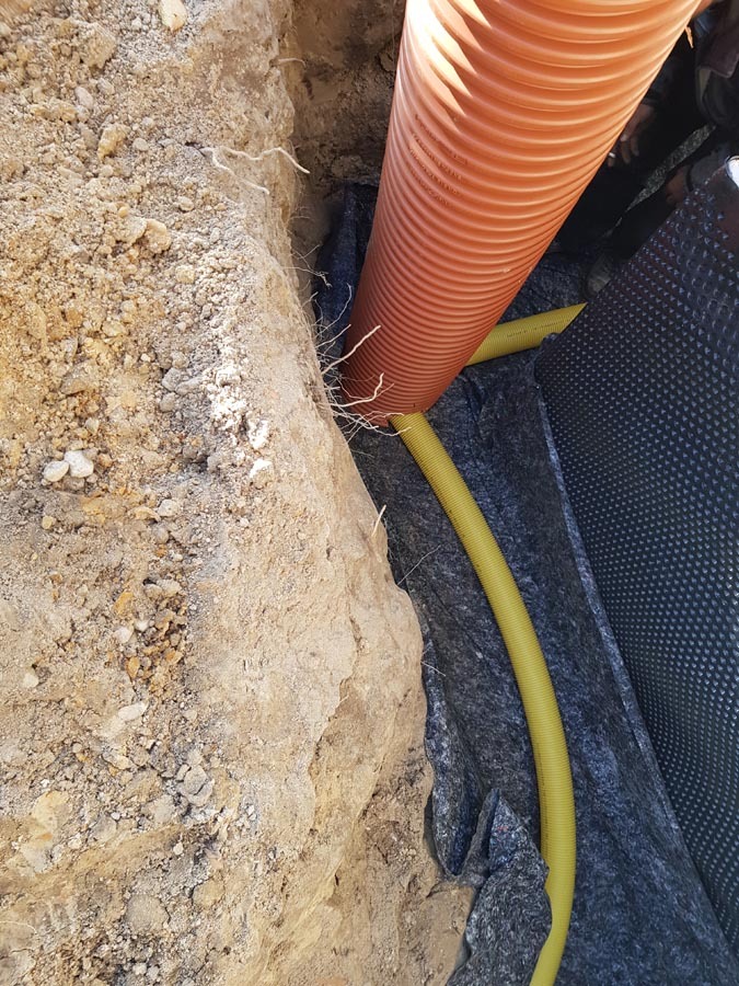 Pomarańczowa rura kanalizacyjna w wykopie, widoczne żółte rury drenarskie i czarna folia izolacyjna na ścianach wykopu, ziemia piaszczysta.