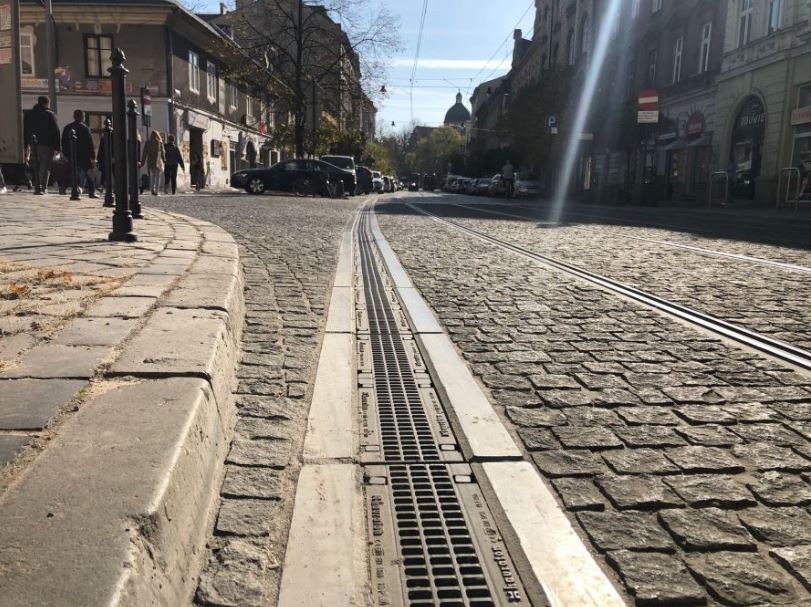 Ulica wybrukowana kostką z systemem odwodnienia liniowego w centralnej części, w tle budynki miejskie i przechodnie.