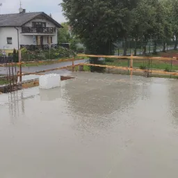 Świeżo wylany, mokry beton na płycie fundamentowej z wystającymi prętami zbrojeniowymi i drewnianym szalunkiem, w tle dom jednorodzinny i boisko sportowe.