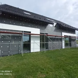 Dom jednorodzinny w trakcie prac związanych z izolacją ścian zewnętrznych, widoczne rusztowania, płyty izolacyjne z charakterystycznymi białymi kołkami mocującymi oraz fragmenty białego tynku.