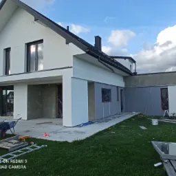 Nowoczesny dom jednorodzinny w trakcie prac elewacyjnych, widoczne fragmenty ocieplenia i tynku, na pierwszym planie taczka i drabina.