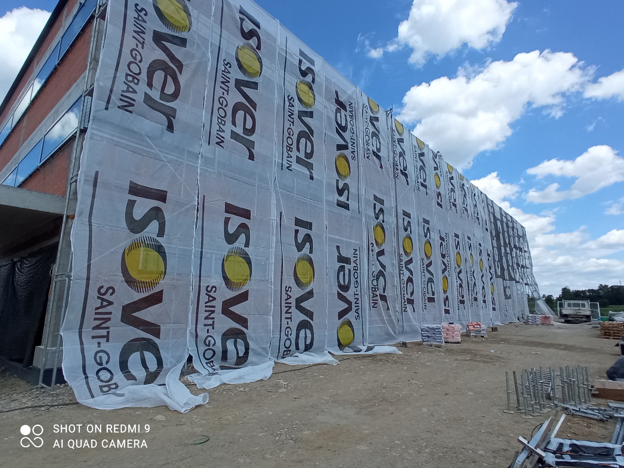 Budynek w trakcie ocieplania, osłonięty folią z logo Isover, widoczne worki z materiałami budowlanymi i rusztowania, błękitne niebo w tle.