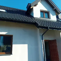 Dom jednorodzinny z nowym, czarnym dachem z dachówki ceramicznej i zamontowanymi rynnami. Widok na fragment elewacji i otwarte drzwi wejściowe.
